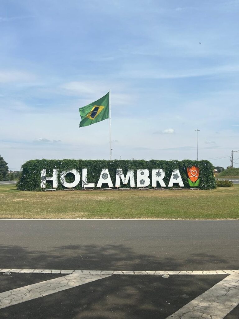 Letreiro escrito Holambra, em frente ao um muro de gramas, com uma bandeira do Brasil hasteado no meio.